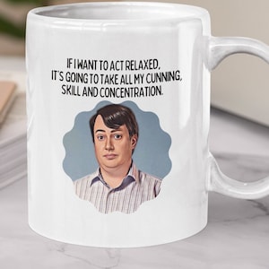 Peut inclure: Une tasse en céramique blanche avec une anse noire. La tasse présente un graphique d'un homme avec une citation : "Si je veux faire semblant d'être détendu, il va falloir que je fasse appel à toute ma ruse, mon talent et ma concentration."