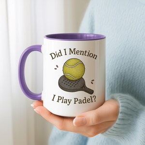 May include: White ceramic mug with a purple handle and interior. The mug features the text "Did I Mention I Play Padel?" along with an illustration of a tennis ball and a padel racket. The mug is held by a person.