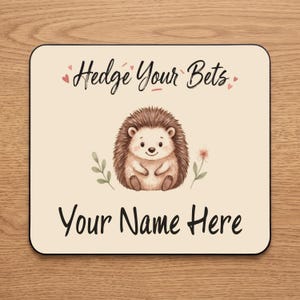 May include: A square coaster with a cream background featuring a brown hedgehog illustration in the centre. The text Hedge Your Bets is printed above the hedgehog with small pink hearts, and the text Your Name Here is printed below in black.