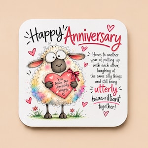 May include: A square coaster featuring a cartoon sheep with rainbow-coloured wool holding a pink heart. The text reads: Happy Anniversary, You Make Me Sheepishly Happy, and Here's to another year of putting up with each other, laughing at the same silly things and still being utterly baaa-rilliant together! Red hearts and small flowers decorate the white background.