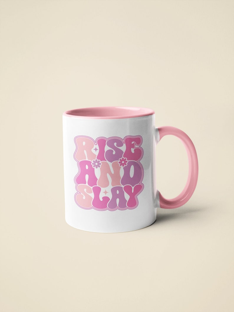 May include: A pink ceramic mug with a pink rim and handle. The mug has a graphic design that says "Rise and Slay" in a retro font with pink, purple, and white colors.