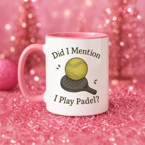 May include: White ceramic mug with a pink handle and interior. The mug features the text "Did I Mention I Play Padel?" along with an illustration of a padel ball and racket. The background is a blurred pink.