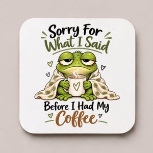 Puede incluir: Un posavasos cuadrado con una rana verde envuelta en una manta de lunares, sosteniendo una taza humeante con un corazón. El texto dice: Sorry For What I Said Before I Had My Coffee. El diseño utiliza letras en negro, verde y marrón.