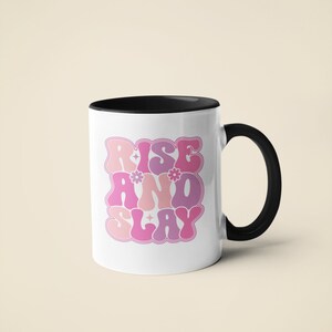 May include: White ceramic mug with a black rim and handle. The mug features a pink and purple graphic that says "Rise and Slay" in a retro font.