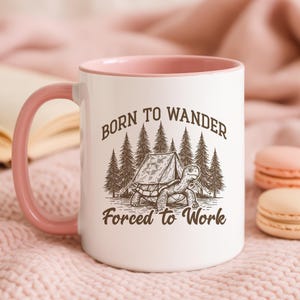 May include: White ceramic mug with a pink handle and interior. The mug features a graphic of a turtle in front of a tent and trees, with the text "Born to Wander Forced to Work" in a brown font.