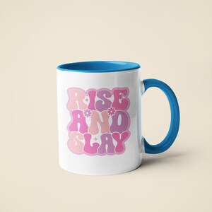 May include: White ceramic mug with a blue rim and handle. The mug has a pink and purple graphic that says "Rise and Slay" with stylized lettering and small flower accents.
