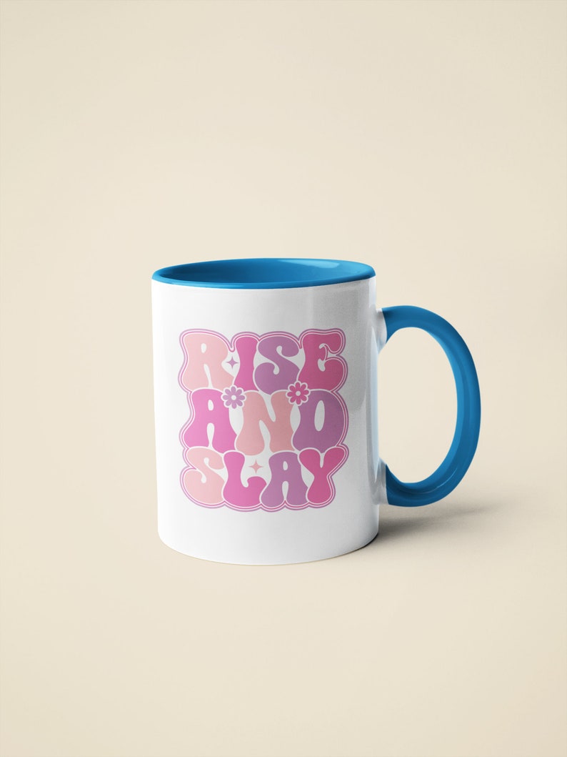 May include: White ceramic mug with a blue rim and handle. The mug has a pink and purple graphic that says "Rise and Slay" with stylized lettering and small flower accents.