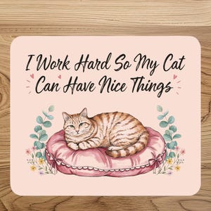 Op de afbeelding: Een rechthoekige lichtroze onderzetter met een bruine cyperse kat die op een rond roze gecapitonneerd kussen ligt. De tekst "I Work Hard So My Cat Can Have Nice Things" staat in zwarte letters boven de kat, geflankeerd door kleine roze hartjes.