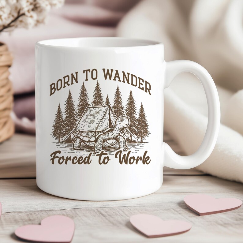 May include: White ceramic mug with a brown graphic of a turtle, tent, and trees. The text on the mug reads "Born to Wander Forced to Work". The mug has a curved handle and is sitting on a light-colored wooden surface.