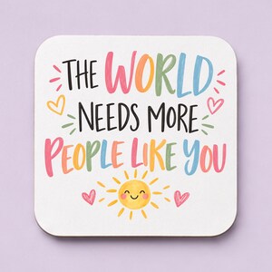 May include: A square white coaster with rounded corners featuring the text THE WORLD NEEDS MORE PEOPLE LIKE YOU in colourful, hand-lettered style font. The design includes small pink hearts and a yellow smiling sun icon at the bottom centre.