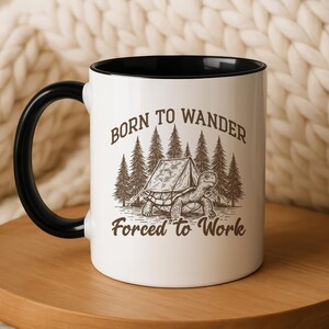 May include: White ceramic mug with a black interior and handle. The mug features a graphic of a turtle with a tent in front of a forest, and the text "Born to Wander Forced to Work" in brown.
