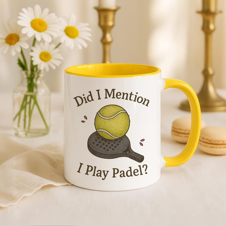 May include: White ceramic mug with a yellow interior and handle. The mug features the text "Did I Mention I Play Padel?" along with a graphic of a padel ball and racket. Daisies in a vase are in the background.