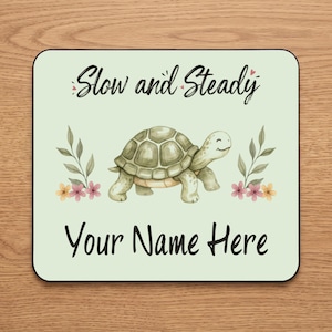 Puede incluir: Una alfombrilla de ratón cuadrada con fondo verde claro que presenta una ilustración central de una tortuga sonriente. El texto "Slow and Steady" aparece sobre la tortuga y "Your Name Here" debajo, flanqueado por pequeñas ramas florales.