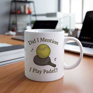May include: White ceramic mug with the text "Did I Mention I Play Padel?" featuring a yellow tennis ball and a padel racket illustration. The mug is on a wooden surface.