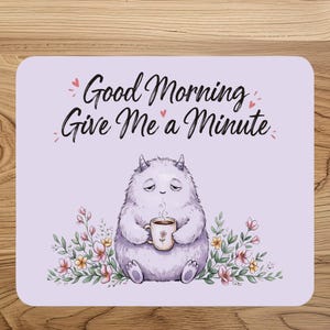 May include: A rectangular light purple mouse pad featuring a sleepy, furry, light purple monster holding a steaming mug. The text Good Morning Give Me a Minute is printed in black above the monster, surrounded by small pink hearts and floral accents.