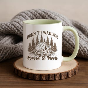 May include: White ceramic mug with a light green interior and handle. The mug features a graphic of a turtle in front of a tent and trees, with the text "BORN TO WANDER Forced to Work". The mug is on a wooden coaster.