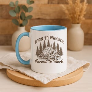 May include: White ceramic mug with a light blue interior and handle. The mug features a graphic of a turtle with a tent and pine trees, and the text "Born to Wander, Forced to Work."