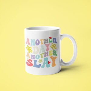 以下が含まれることがあります： 花とハートがあしらわれたカラフルなレトロな文字で「Another Day Another Slay」と書かれた白いセラミックマグカップ