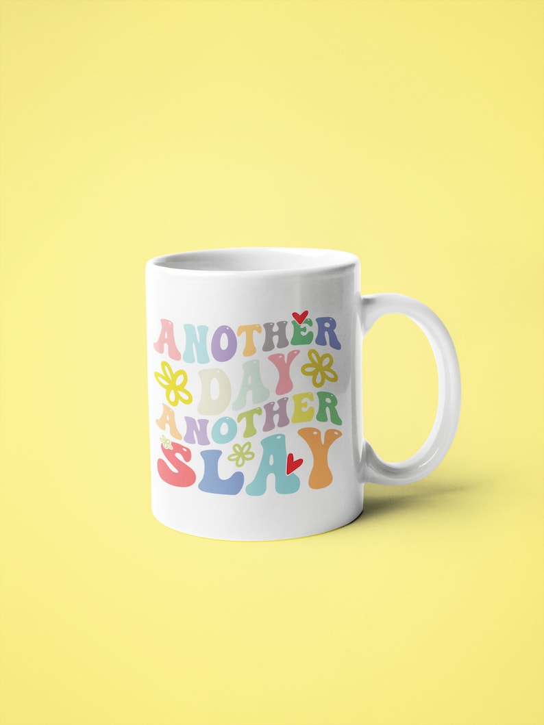 以下が含まれることがあります： 花とハートがあしらわれたカラフルなレトロな文字で「Another Day Another Slay」と書かれた白いセラミックマグカップ