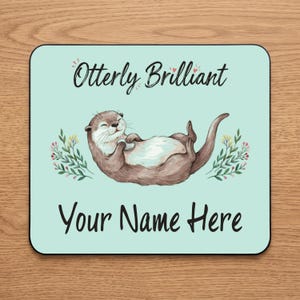 May include: A square mouse pad with a light mint green background featuring a brown otter illustration, floral accents, and the text "Otterly Brilliant" at the top and "Your Name Here" at the bottom in a black cursive and print font.