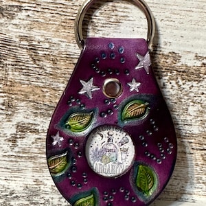 May include: Purple leather keychain with a round, clear window featuring a drawing of a tequila bottle and margarita glass. The keychain is decorated with green leaves, white stars, and black dots.