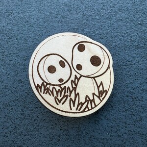 May include: A wooden circle with a laser-cut design of two mushrooms with black spots and a green grass border.