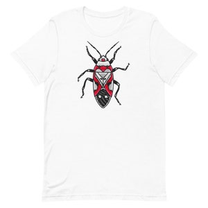 Pode incluir: Uma camiseta branca com uma ilustração de um inseto vermelho, preto e branco. O inseto tem um corpo longo e fino com seis patas e um par de antenas.