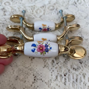 May include: Set of four cabinet pulls with a gold finish and white ceramic handles. Each handle is decorated with a floral design in pink, blue, and yellow. The pulls are shown with their mounting screws.