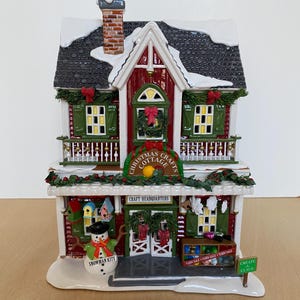 Może przedstawiać: Figurka Christmas Cottage z czerwoną i białą fasadą, zielonymi okiennicami i czarnym dachem pokrytym śniegiem. Znak głosi "Christmas Cottage" i "Craft Headquarters". Figurka bałwana i znak z napisem "Snowman Kits" znajdują się z przodu.