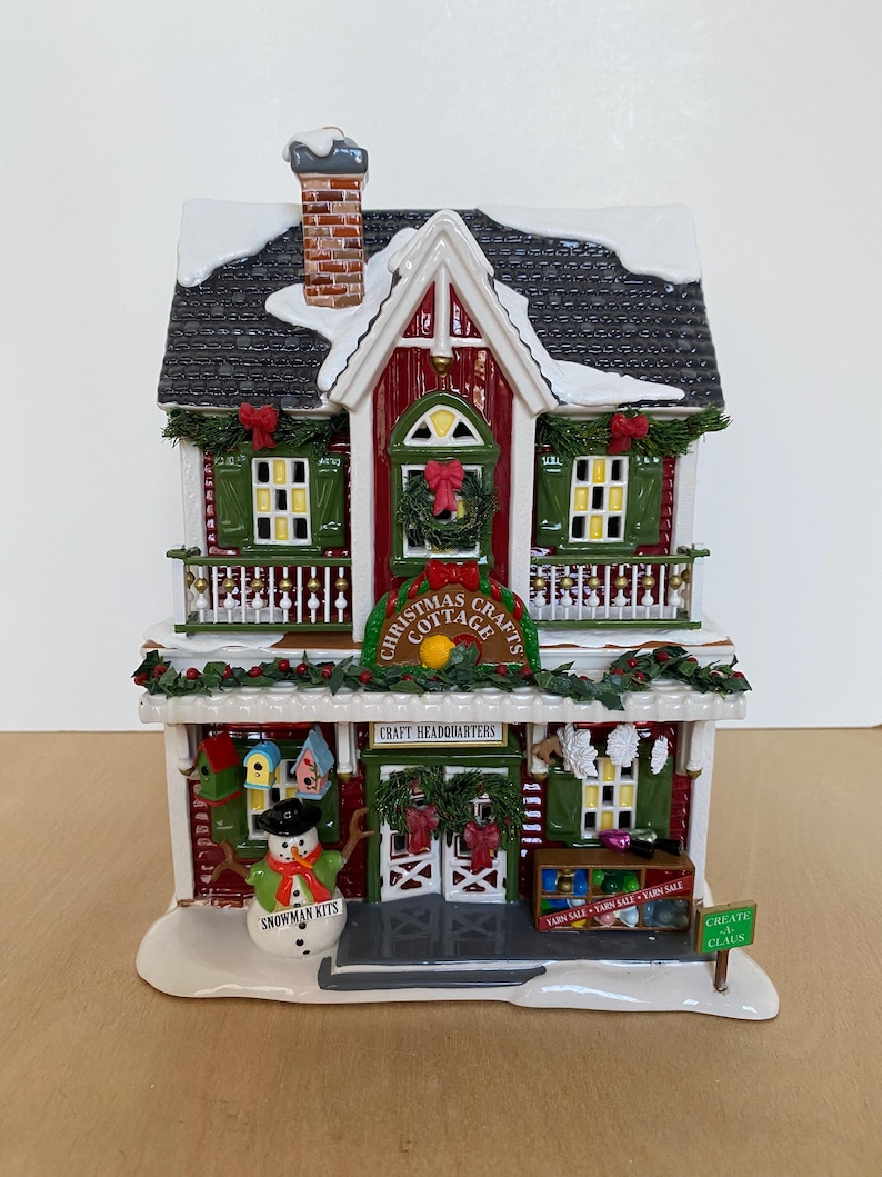 Może przedstawiać: Figurka Christmas Cottage z czerwoną i białą fasadą, zielonymi okiennicami i czarnym dachem pokrytym śniegiem. Znak głosi "Christmas Cottage" i "Craft Headquarters". Figurka bałwana i znak z napisem "Snowman Kits" znajdują się z przodu.