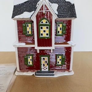 Może przedstawiać: Czerwona ceramiczna figurka domu z czarnym dachem i białymi akcentami śnieżnymi. Dom ma zielone okiennice i ż&oacute;łto oświetlone okna. Widoczny jest r&oacute;wnież mały komin i drzwi wejściowe. Przedmiot dekoracyjny.