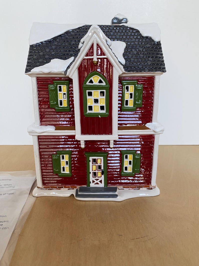Może przedstawiać: Czerwona ceramiczna figurka domu z czarnym dachem i białymi akcentami śnieżnymi. Dom ma zielone okiennice i ż&oacute;łto oświetlone okna. Widoczny jest r&oacute;wnież mały komin i drzwi wejściowe. Przedmiot dekoracyjny.