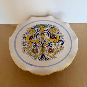 May include: A round ceramic cake stand with a scalloped edge, featuring a cream-colored surface decorated with a central motif of two yellow and blue mythical creatures flanking a floral design, all enclosed by a thin light blue circular border.