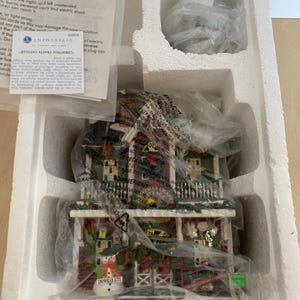 Może przedstawiać: Figurka Christmas Crafts Cottage, zapakowana w styropianowe pudełko. Domek ma czerwony dach, zielone obramowania i figurkę bałwana. Opakowanie zawiera instrukcje bezpieczeństwa i etykietę Department 56.