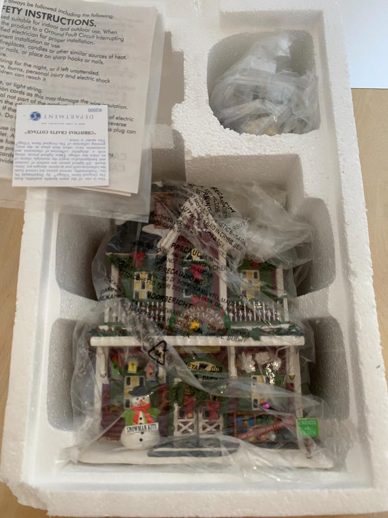 Może przedstawiać: Figurka Christmas Crafts Cottage, zapakowana w styropianowe pudełko. Domek ma czerwony dach, zielone obramowania i figurkę bałwana. Opakowanie zawiera instrukcje bezpieczeństwa i etykietę Department 56.