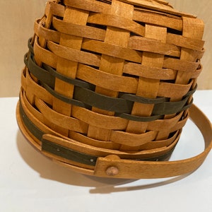 May include: A woven basket with a handle. The basket is made of light brown wood and has a dark green band around the middle. The handle is also made of wood and is attached to the basket with a metal rivet.