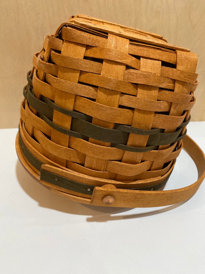 May include: A woven basket with a handle. The basket is made of light brown wood and has a dark green band around the middle. The handle is also made of wood and is attached to the basket with a metal rivet.