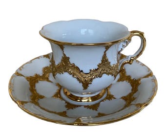 May include: A white porcelain teacup and saucer set featuring a scalloped rim and an ornate, raised gold filigree pattern. The cup has a curved handle with gold detailing, and the saucer displays a repeating gold lattice design.