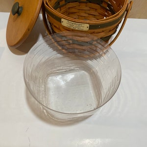 May include: A brown woven basket with a wooden lid and handle. The basket has a green stripe and a metal tag that reads "Longaberger Christmas Collection Jingle Bell Basket". A clear plastic container is in front.