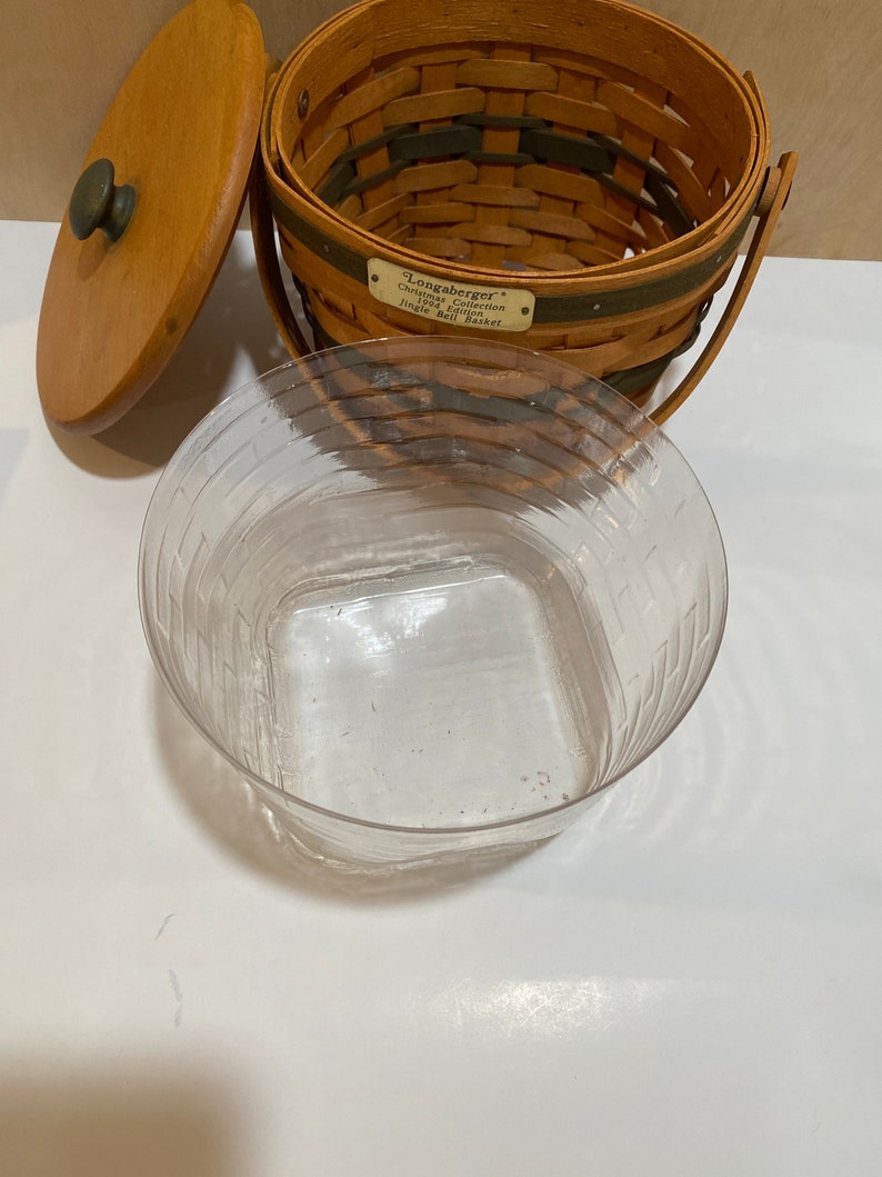 May include: A brown woven basket with a wooden lid and handle. The basket has a green stripe and a metal tag that reads "Longaberger Christmas Collection Jingle Bell Basket". A clear plastic container is in front.