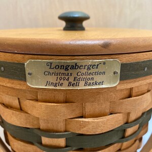 May include: A woven basket with a wooden lid and handle. The basket is made of light brown and green woven material. A metal tag reads "Longaberger Christmas Collection 1994 Edition Jingle Bell Basket."