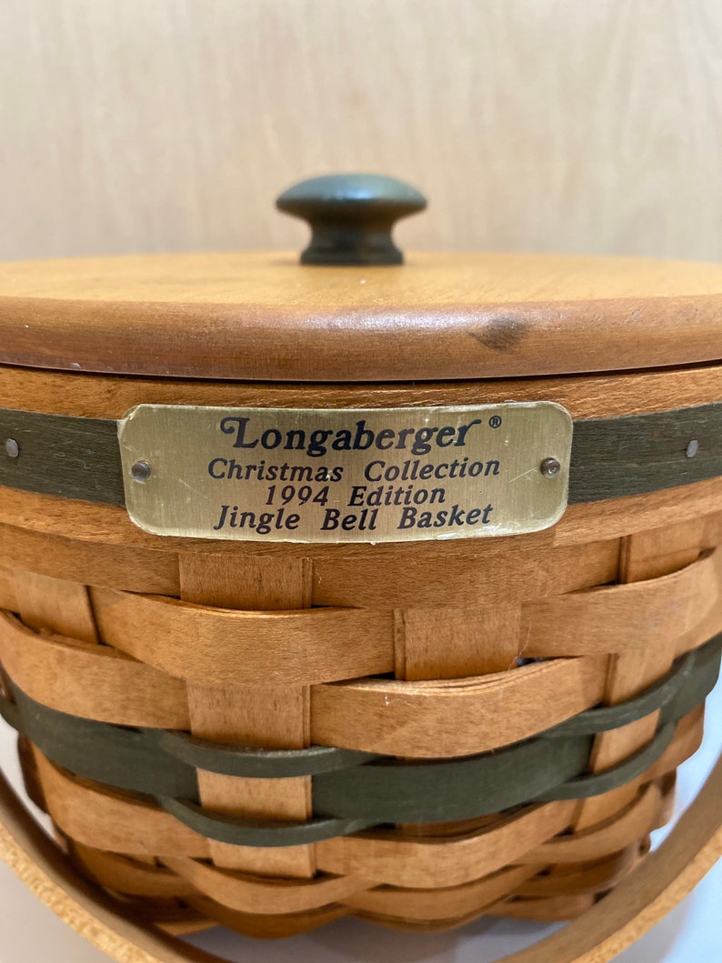 May include: A woven basket with a wooden lid and handle. The basket is made of light brown and green woven material. A metal tag reads "Longaberger Christmas Collection 1994 Edition Jingle Bell Basket."