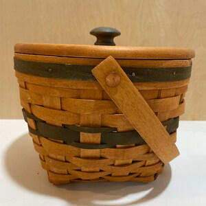 May include: A lidded woven basket with a handle. The basket is made of light brown woven material with dark green accents. The lid has a dark green knob. The handle is made of light brown wood and is attached with a copper rivet.