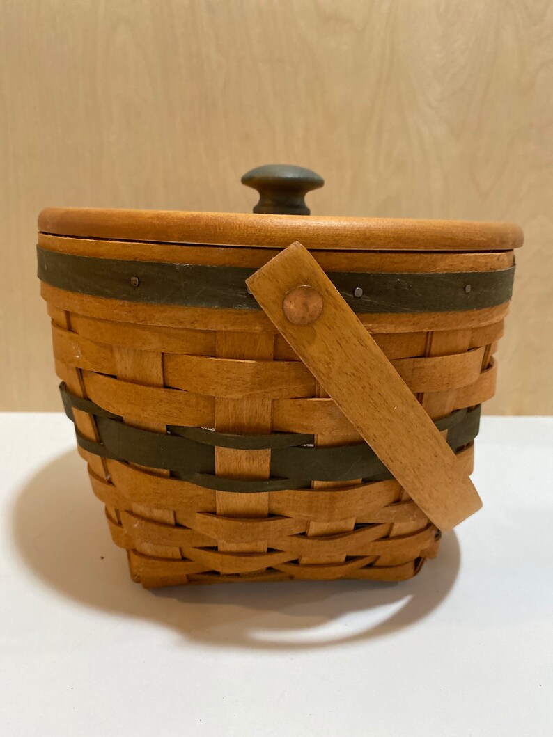 May include: A lidded woven basket with a handle. The basket is made of light brown woven material with dark green accents. The lid has a dark green knob. The handle is made of light brown wood and is attached with a copper rivet.