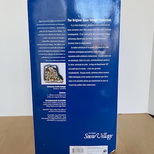 Może przedstawiać: Niebieskie pudełko dla "The Original Snow Village Collection" zawiera tekst i obrazy. Pudełko zawiera informacje o produkcie, w tym "Christmas Crafts Cottage" i logo Department 56. Pudełko jest przeznaczone do ceramicznych element&oacute;w wioski.