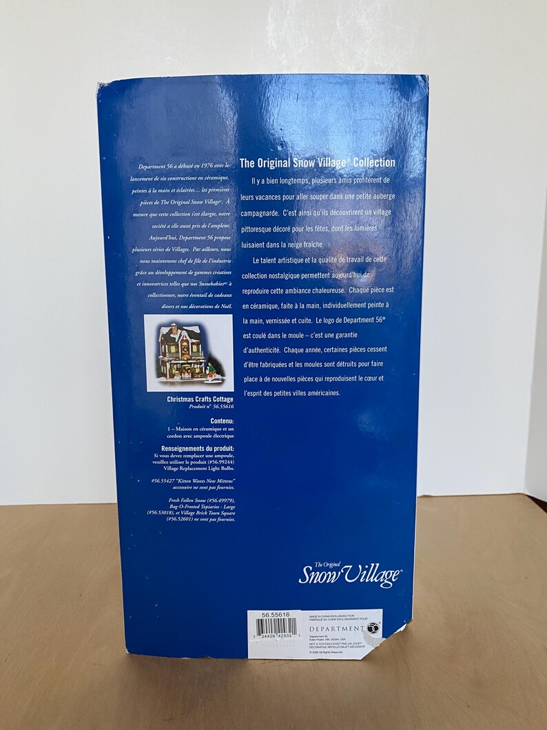 Może przedstawiać: Niebieskie pudełko dla "The Original Snow Village Collection" zawiera tekst i obrazy. Pudełko zawiera informacje o produkcie, w tym "Christmas Crafts Cottage" i logo Department 56. Pudełko jest przeznaczone do ceramicznych element&oacute;w wioski.