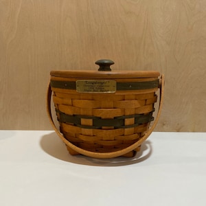 May include: A Longaberger Christmas Collection 1994 Edition Jingle Bell Basket. The basket is made of woven brown material with dark green accents. It has a wooden lid with a dark knob and a swing handle. A brass plate displays the collection name.