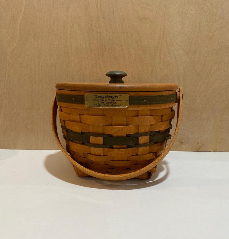 May include: A Longaberger Christmas Collection 1994 Edition Jingle Bell Basket. The basket is made of woven brown material with dark green accents. It has a wooden lid with a dark knob and a swing handle. A brass plate displays the collection name.