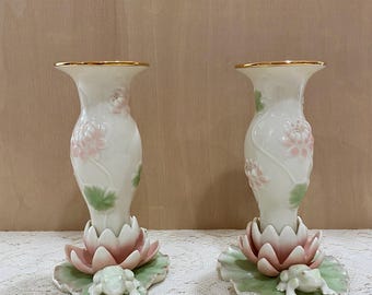 May include: A pair of white porcelain candle holders with a gold rim. The holders are decorated with pink and green flowers and a frog on a leaf base.