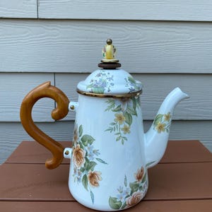 May include: White enamel teapot with a floral design featuring orange, yellow, and blue flowers. The teapot has a wooden handle and a lid with a decorative knob. The rim is trimmed with gold.